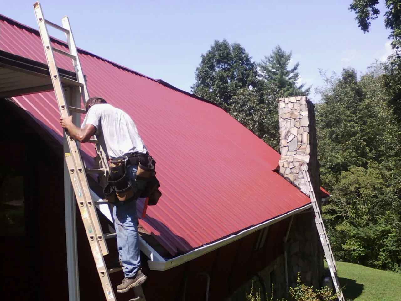 Roofer on a ladder beside a red metal roof and stone chimney — Soffit & Fascia Repair by Roofing Brothers in Chippewa Lake